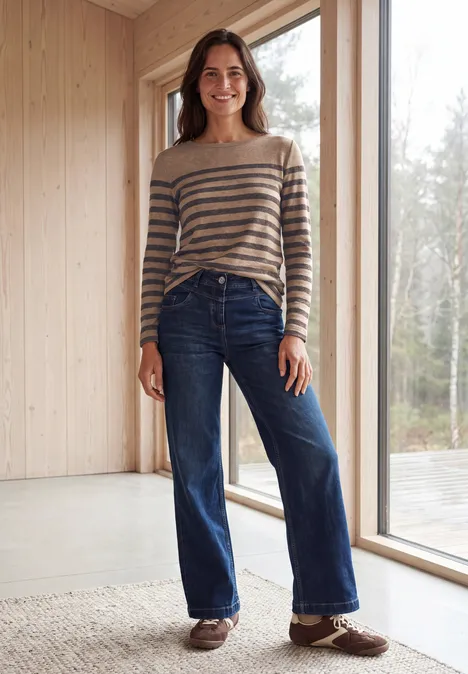 Frau mit gestreiftem Pullover und weiter Jeans steht vor großem Fenster in hellem Raum.