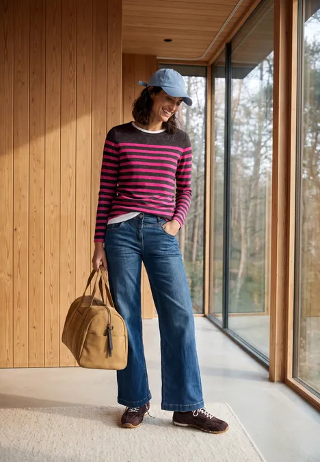 T-shirt rayé à manches longues et col bateau truffle brown melange Frau in rosa-blau gestreiftem Pullover weitem Jeans, blauer Kappe und Tasche steht in hellem Holzkorridor mit Fenstern.