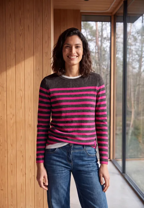 T-shirt rayé à manches longues et col bateau truffle brown melange Frau steht in modernem Holzinterieur, trägt gestreiften Pullover und Jeans, lächelt in die Kamera.