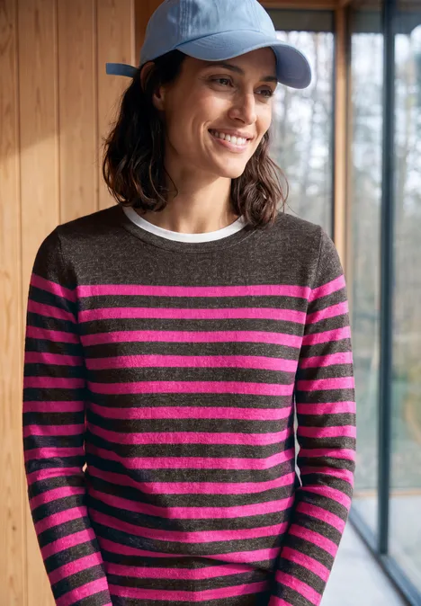 T-shirt rayé à manches longues et col bateau truffle brown melange Frau mit blauem Baseball-Cap und grau-pink gestreiftem Pullover, die lächelnd vor einem Fenster steht.