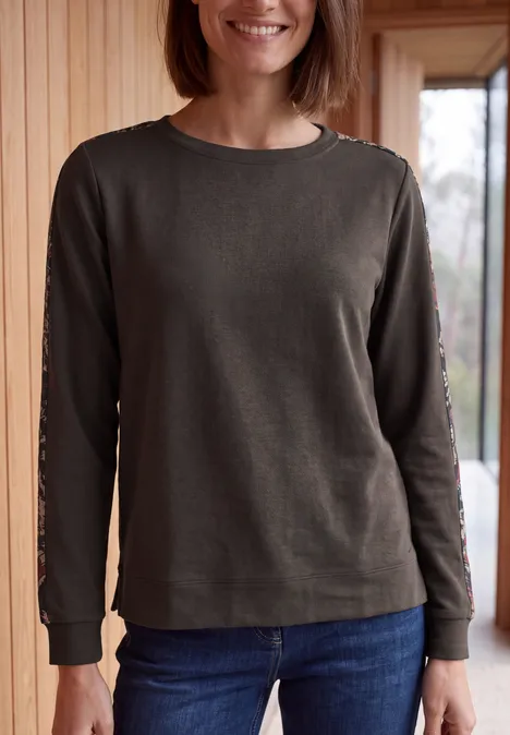 Shirt mit Boat Neck und Print-Tape truffle brown Frau mit grünem Langarmshirt und Jeans steht lächelnd vor einer Holzpaneelwand.