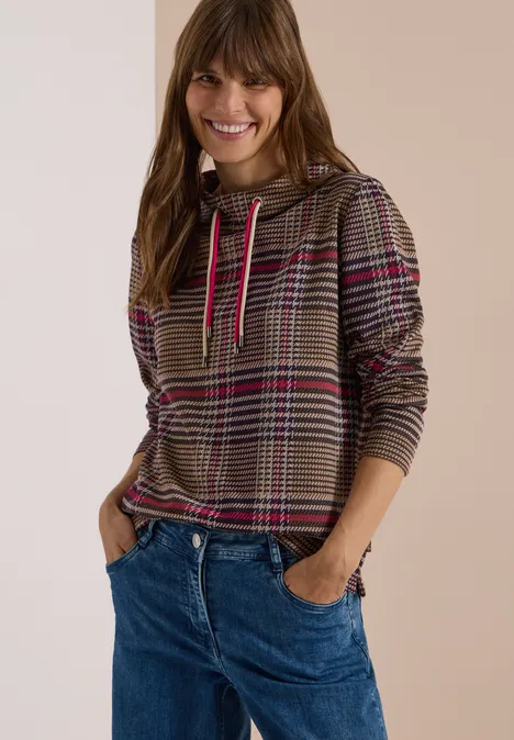 Glimlachende vrouw in geruit hoodie met roze strepen en blauwe jeans, handen in de zakken.
