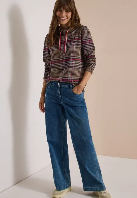 Vrouw lacht in een geruite hoodie en wijde jeans tegen een neutrale achtergrond.