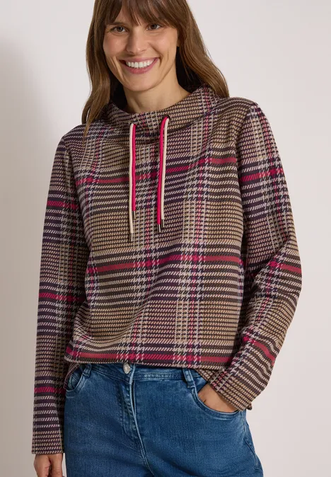 Lachende vrouw in bruin geruite hoodie met roze strepen en blauwe spijkerbroek, witte achtergrond.