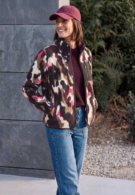 Fleecejas met lange mouwen en print pink flash Frau mit Camouflage-Jacke, Jeans und Cap lächelt draußen vor einer Steinwand und Büschen.