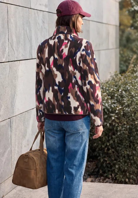 Fleecejas met lange mouwen en print pink flash Frau mit roter Mütze, gemusterter Jacke und Jeans trägt eine braune Tasche vor Mauer und Sträuchern.