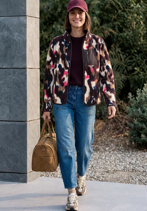 Fleecejas met lange mouwen en print pink flash Frau mit bunter Jacke, Jeans, Sneakers und Baseballcap trägt Tasche und spaziert vor einem Haus.