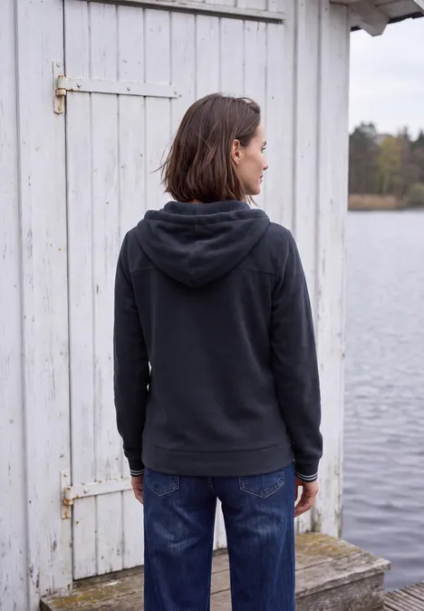 Langarm Fleece Jacke mit Kapuze urban dark blue Rückansicht einer Person in dunkelgrauer Kapuzenjacke vor einem weißen Holzhaus am Wasser.