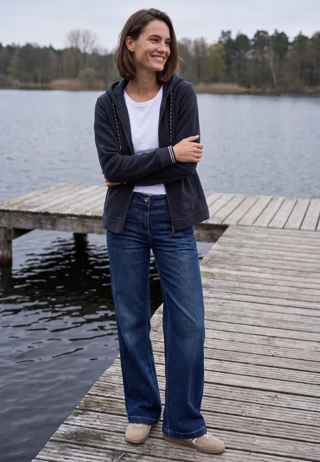 Langarm Fleece Jacke mit Kapuze urban dark blue Frau lächelnd in Jeans und Cardigan steht auf einem Steg am ruhigen See mit Wald im Hintergrund.