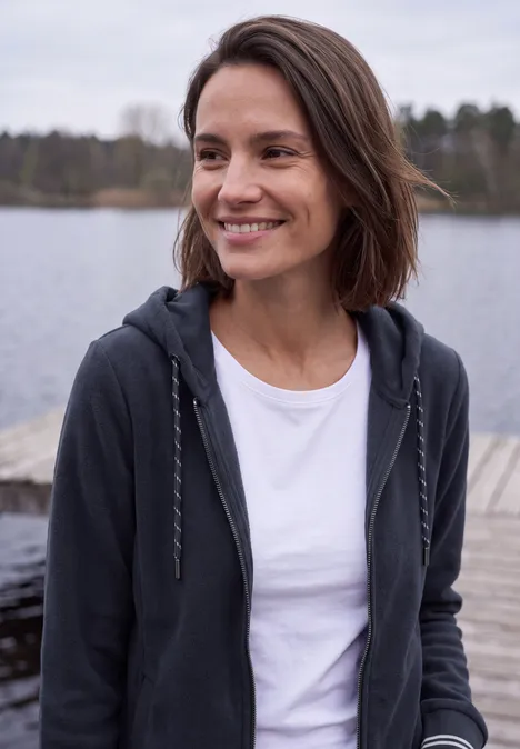 Langarm Fleece Jacke mit Kapuze urban dark blue Eine lächelnde Frau im weißen T-Shirt und Hoodie steht auf einem Steg am See, im Hintergrund Wald.