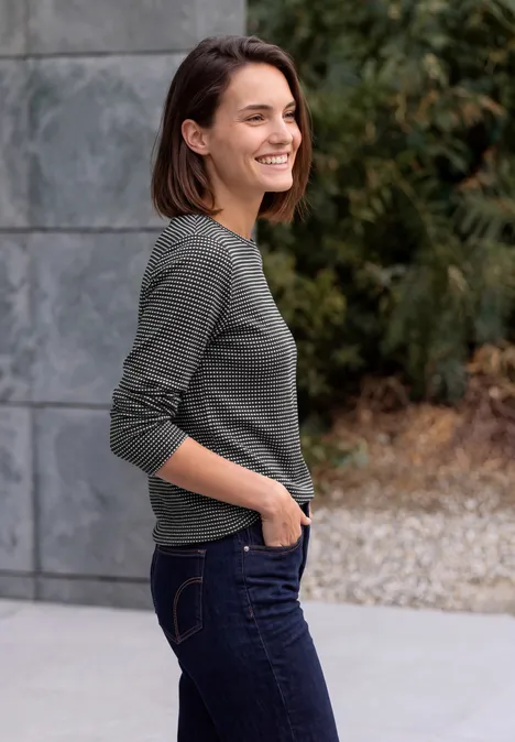 Eine Frau steht draußen in einem grauen Langarmshirt und Jeans, lächelnd mit der Hand in der Tasche.