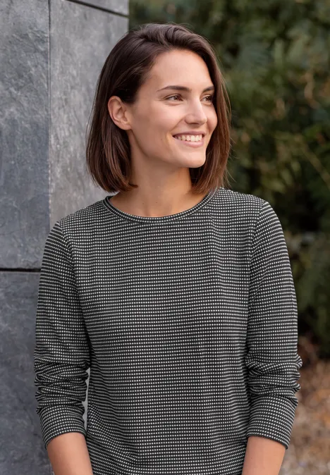 Frau mit braunen Haaren lächelt im Freien, trägt graues, gestreiftes Langarmshirt vor einer Steinmauer.