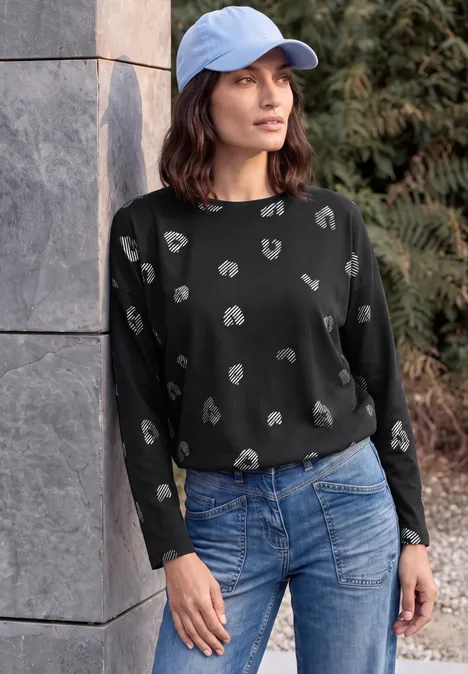 Langarm Shirt mit Boat Neck und Leo-Muster Black Eine Frau lehnt lässig an einer Mauer, trägt einen schwarzen Pullover mit weißem Muster, Jeans und eine blaue Kappe.