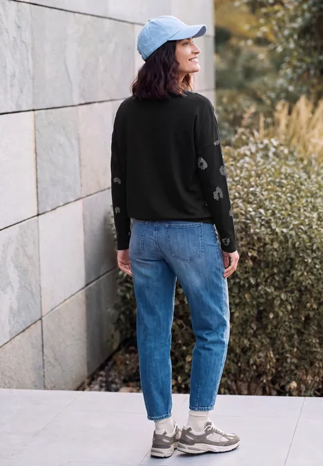 Langarm Shirt mit Boat Neck und Leo-Muster Black Junge Frau in Jeans, schwarzem Pullover und Cap steht vor einer Steinmauer neben Büschen.