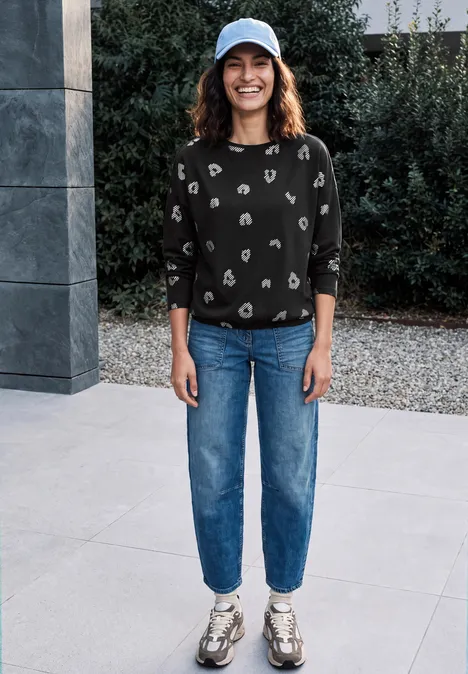 Langarm Shirt mit Boat Neck und Leo-Muster Black Frau steht lächelnd draußen vor modernem Bauwerk, trägt Cap, schwarz gemusterten Pullover, Jeans und Sneaker.