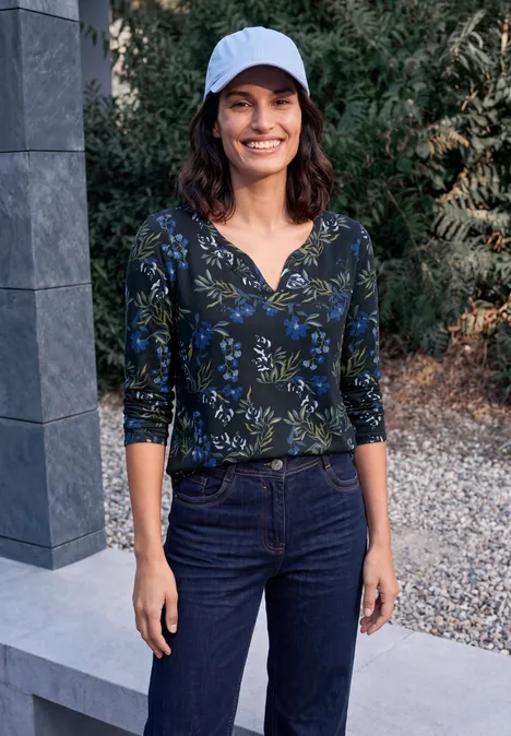 Une femme en casquette blanche, top à fleurs bleu-vert et jean, debout à l'extérieur, souriante.