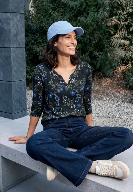 Femme casquette bleue assise jambes croisées sur banc en pierre, souriante, vêtue d’un haut fleuri, jean et baskets blanches.