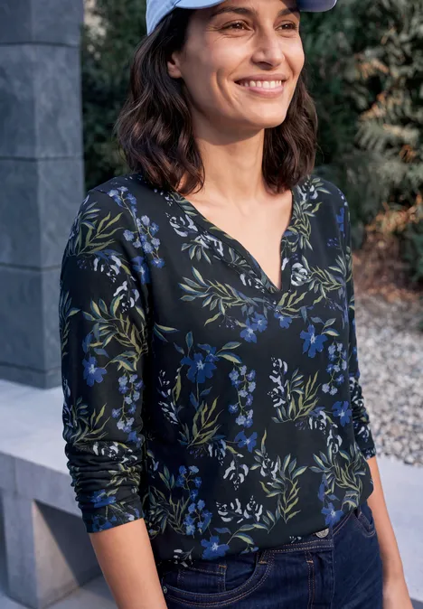 Une femme souriante en haut à fleurs bleu-vert et casquette se tient dehors devant un mur en pierre.