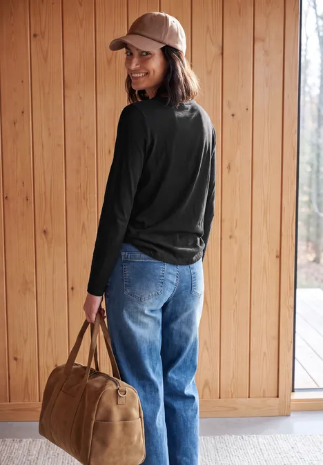 Une femme en jean, haut noir à manches longues et casquette, porte un sac et sourit devant un mur en bois.