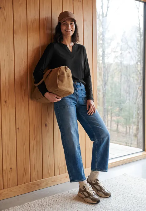 Femme en jean, veste sombre et casquette beige, sac à main, posant contre un mur en bois près d'une fenêtre.