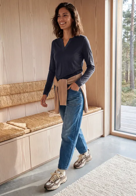Femme en jean, haut bleu marine et baskets se promenant à l'intérieur d'une pièce moderne et lumineuse près d'un banc en bois.