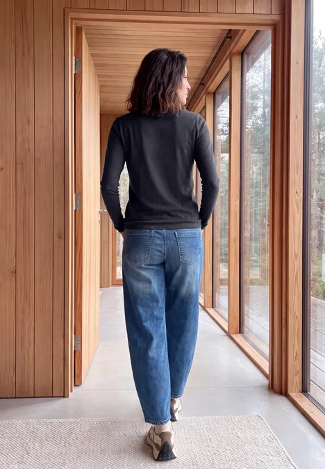 Une personne debout dans un couloir en bois lumineux, vêtue d’un pull et d’un jean, regarde à travers les grandes fenêtres.