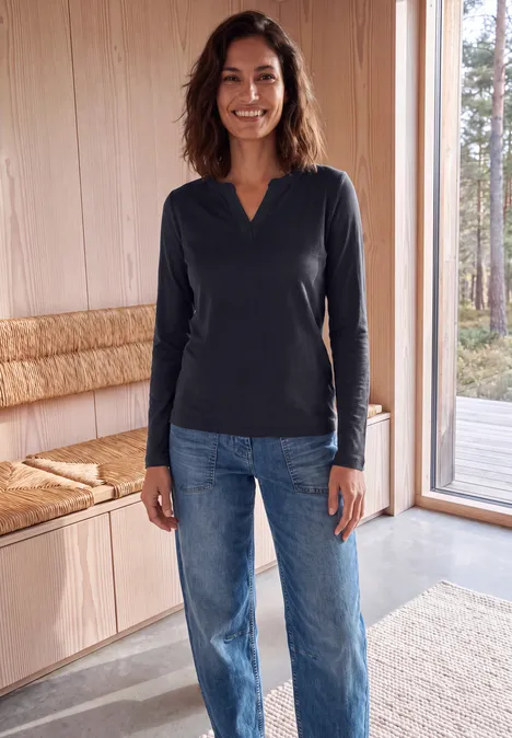 Femme souriante en jean et haut à manches longues foncé, debout près d'une fenêtre dans une pièce chaleureuse.