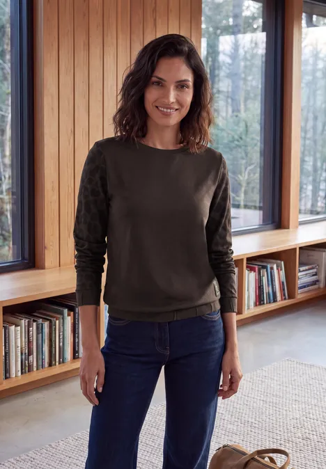 T-shirt à col rond et manches léopard truffle brown Eine lächelnde Frau in grünem Pullover und Jeans steht in einem hellen Raum vor Bücherregalen und Fenstern.