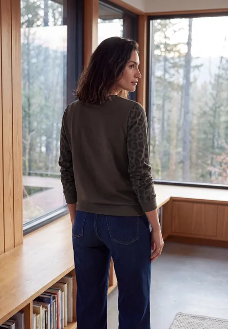 T-shirt à col rond et manches léopard truffle brown Frau steht rücklings in einem hellen Holzraum, lehnt an Sitzbank, trägt Jeans und grünen Pullover.