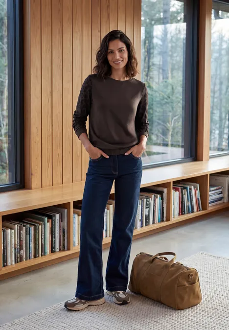 T-shirt à col rond et manches léopard truffle brown Eine Frau in Jeans, Pullover und Sneakers steht in einem hellen Bücherraum mit Reisetasche.