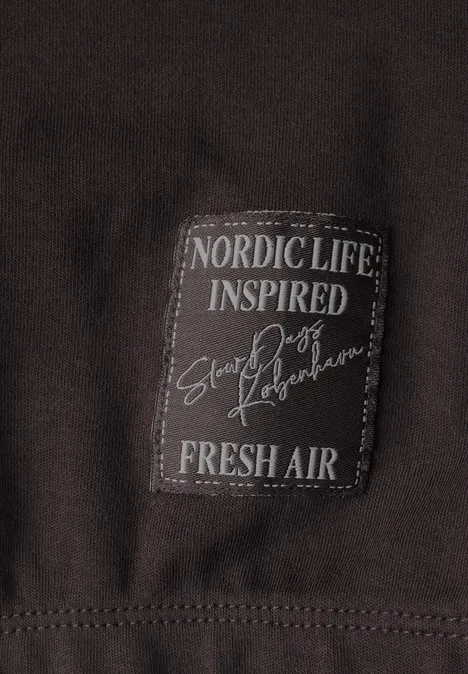 T-shirt à col rond et manches léopard truffle brown Nahaufnahme eines dunkelbraunen Kleidungsstücks mit aufgenähter Text-Plakette: ‚NORDIC LIFE INSPIRED Snow Poets FRESH AIR‘.