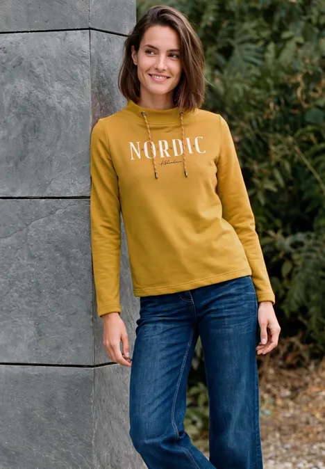 Eine Frau lehnt lächelnd an einer grauen Säule, trägt einen senfgelben Pullover mit 'NORDIC' und blaue Jeans.
