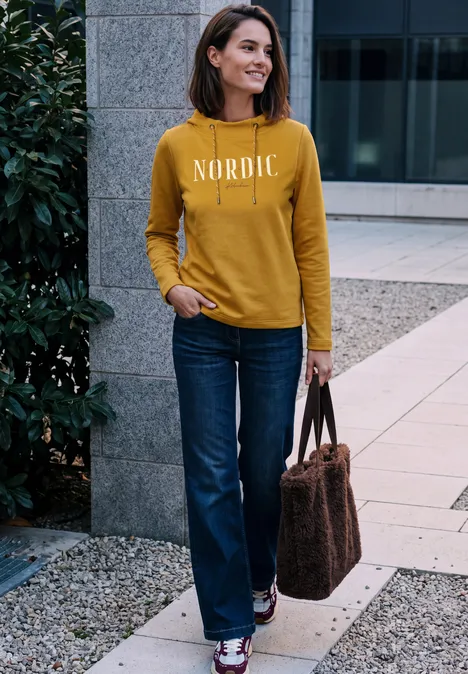 Frau im senfgelben Pullover mit Schriftzug 'NORDIC', Jeans und Tasche steht lächelnd vor moderner Fassade.