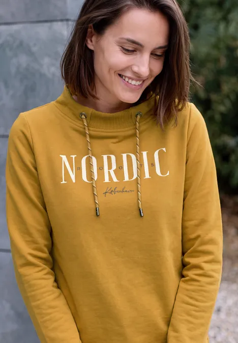 Junge Frau in senfgelbem Hoodie mit Aufschrift 'NORDIC', lächelnd und nach unten blickend.