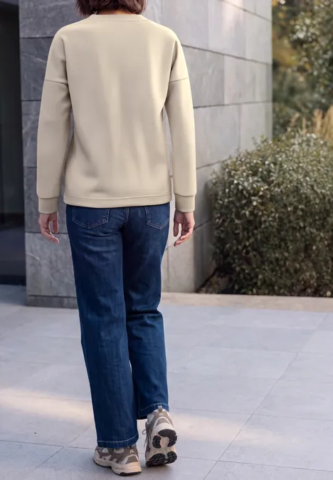 T-shirt à manches longues, col rond et texture côtelée pearl beige Person von hinten in beigem Pullover, blauen Jeans und Sneakers auf Pflaster neben Mauer und Sträuchern.