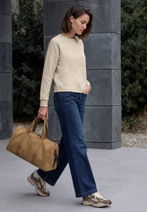 T-shirt à manches longues, col rond et texture côtelée pearl beige Frau mit kurzem Haar in beigem Pullover, weitem Jeans, Sneakern und großer brauner Tasche vor Steinwand.