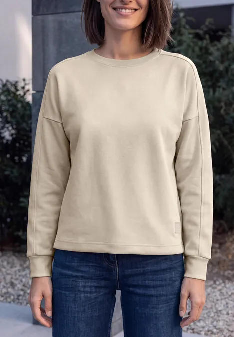 T-shirt à manches longues, col rond et texture côtelée pearl beige Frau in beigem Sweatshirt und blauen Jeans steht vor uns, lächelnd.