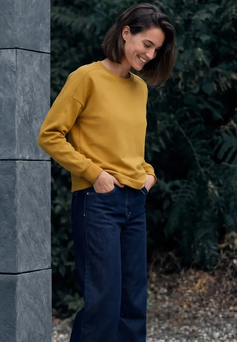 T-shirt à manches longues, col rond et texture côtelée golden honey Eine lächelnde Frau in gelbem Pullover und Jeans lehnt an einer Steinwand vor grünen Pflanzen.