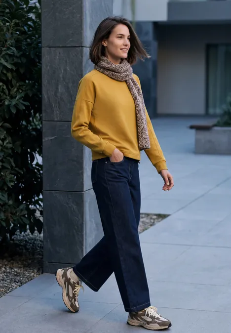 T-shirt à manches longues, col rond et texture côtelée golden honey Frau in gelbem Pullover, Schal und dunkelblauer Hose spaziert lächelnd vor modernem Gebäude.