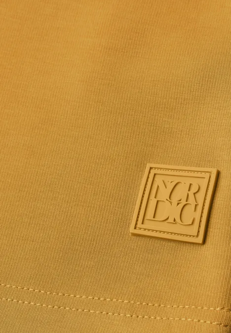 T-shirt à manches longues, col rond et texture côtelée golden honey Senfgelber Stoff mit quadratischem Y.R.C.C.-Logo-Patch in Nahaufnahme.