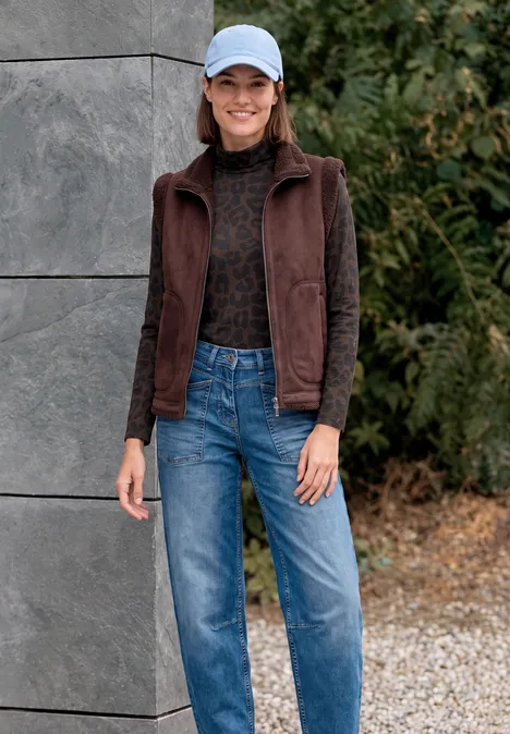 T-shirt à manches longues, col roulé et motif léopard truffle brown Eine lächelnde Frau mit hellblauer Mütze, brauner Weste, schwarzem Pullover und Jeans vor Betonwand und Grün.