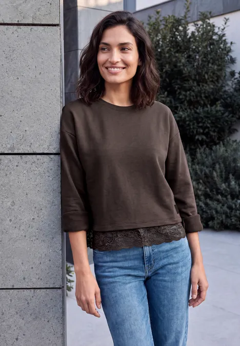 Une femme souriante en pull marron et jean bleu appuyée contre un mur à l'extérieur.