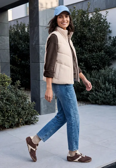 Femme en doudoune sans manches beige, jeans et baskets, souriante, marchant devant un bâtiment et des buissons.