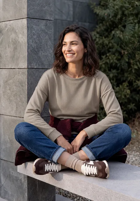 T-shirt à manches longues avec motif en chevrons pearl beige Eine Frau sitzt im Freien auf einer Betonbank, lächelt und trägt Pullover, Jeans und Sneakers.