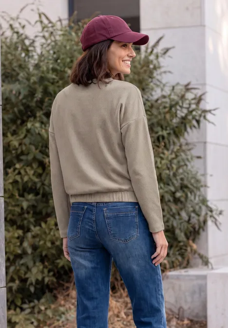 T-shirt à manches longues avec motif en chevrons pearl beige Frau mit roter Baseballmütze, olivgrünem Pullover und Jeans steht im Freien und lächelt über die Schulter.