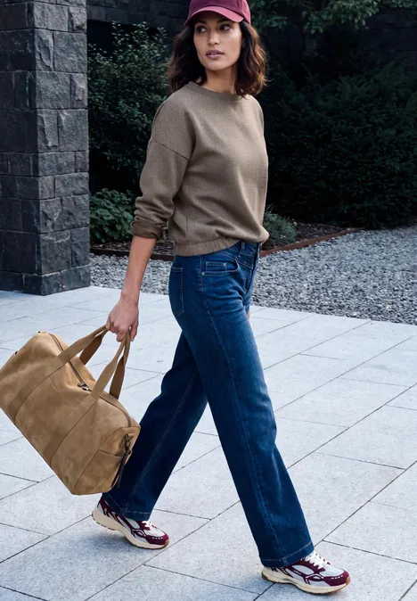 Frau in Freizeitkleidung mit Jeans, Pullover und Tasche schlendert auf gepflastertem Weg.