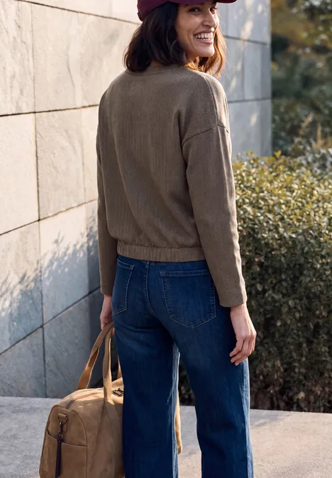 Frau im Jeans-Outfit, Jacke und Cap, trägt Tasche, steht an Wand im Freien.