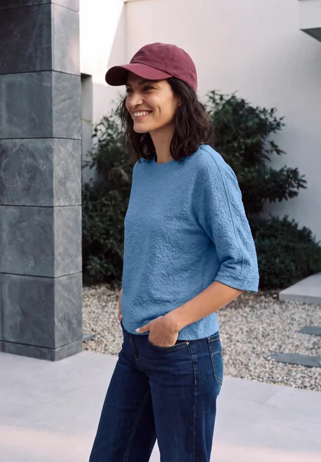 3/4-Arm Shirt mit Rundhals und Leo-Muster north blue Frau mit roter Baseballkappe, blauem Shirt und Jeans steht lächelnd draußen vor modernem Gebäude.