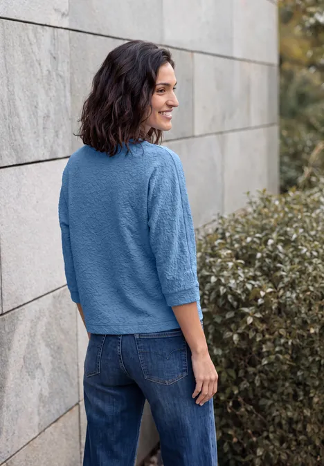 3/4-Arm Shirt mit Rundhals und Leo-Muster north blue Eine lächelnde Frau in blauem Pullover und Jeans lehnt an einer Mauer im Freien, im Profil.
