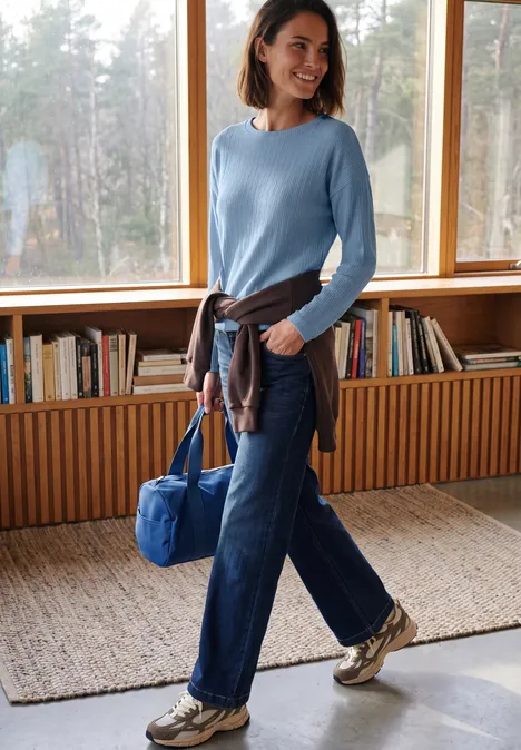 Ottoman Shirt mit Boat Neck cosmopolitan blue Frau in hellem Raum trägt hellblauen Pullover, Jeans und Sneakers, hält blaue Tasche vor Bücherregal.